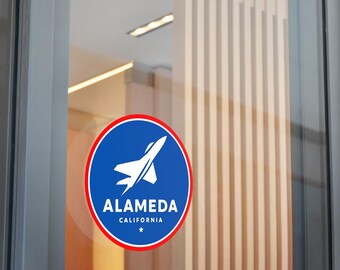 May include: A round sticker with a blue background and a white jet plane graphic. The sticker has a red border and text that reads "ALAMEDA CALIFORNIA" with a star below. The sticker is affixed to a glass door.