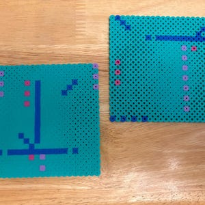 May include: Two teal square pixel art designs. One features a vertical blue line with a horizontal blue line at the bottom, and pink and purple dots. The other has a blue design and pink dots. The squares are made of small, round beads.