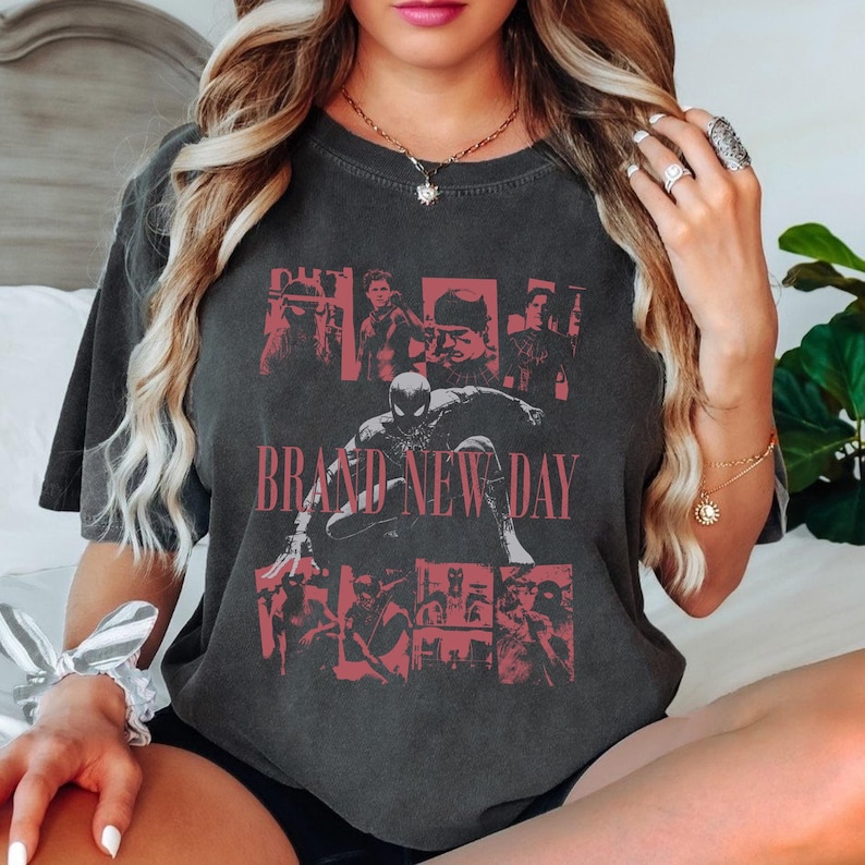 May include: Dark gray t-shirt featuring a red graphic of Spider-Man and other characters, with the text "BRAND NEW DAY". The design is a collage of images. The shirt has short sleeves and a crew neck.