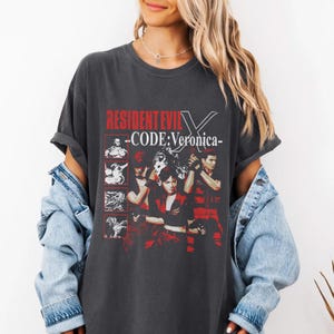 May include: Dark gray t-shirt featuring the text "RESIDENT EVIL - CODE: Veronica-" in red and white. The shirt has a graphic design with images of characters from the video game. The shirt is a relaxed fit.