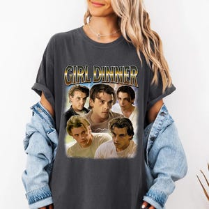 May include: Dark gray t-shirt featuring a graphic with the text "GIRL DINNER" in gold lettering above a collage of five male faces. The shirt has short sleeves and a relaxed fit.