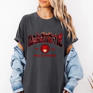 May include: Dark gray t-shirt featuring the word "DAREDEVIL" in a red, textured font with a group of characters. Below the word is a red circle with a masked face and the text "EST. 1965 HELL'S KITCHEN".
