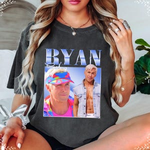 May include: Dark gray t-shirt featuring a graphic of a man with the name "RYAN" in a blue, stylized font. The graphic includes two images of the man, one wearing a visor and the other a denim vest. The text "Ken" is visible.