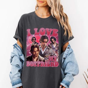 May include: Dark gray t-shirt with a pink graphic that says "I LOVE MY BOYFRIEND." The graphic features a heart and multiple images of a man. The shirt has short sleeves and a crew neck.