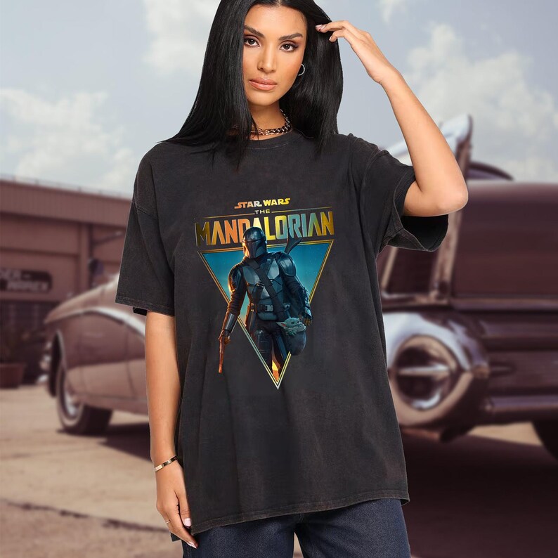 Op de afbeelding: Zwart T-shirt met een afbeelding van The Mandalorian uit Star Wars. Het ontwerp is geplaatst in een blauwe en gouden driehoek, met het Star Wars-logo erboven. Het shirt heeft korte mouwen en een relaxte pasvorm.