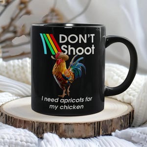 May include: Black ceramic mug featuring a colorful rooster wearing a cowboy hat. The mug has the text "DON'T Shoot" above the rooster and "I need apricots for my chicken" below. The mug has a black handle.