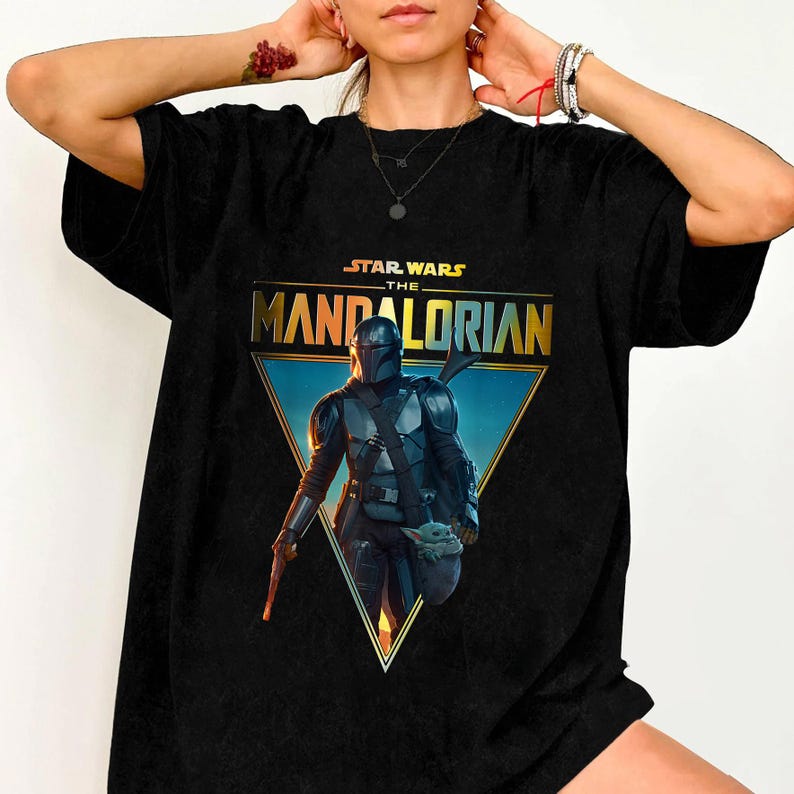 Op de afbeelding: Zwart T-shirt met een afbeelding uit de Star Wars-serie, "The Mandalorian". Het ontwerp bevat de titel van de show in gouden letters, een driehoekige achtergrond en een afbeelding van The Mandalorian en Grogu.