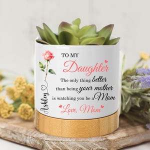 May include: A white ceramic planter with a bamboo base, holding a succulent plant. The planter is decorated with a pink rose and the text "To My Daughter". A heartfelt gift for a mother.