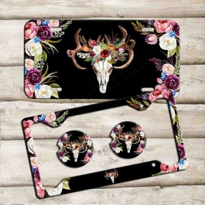 May include: A black and white deer skull with a floral wreath on its antlers. The skull is surrounded by a floral border with pink, white, and purple flowers. The image is for a set of car accessories, including a license plate frame and two drink coasters.