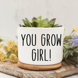 May include: White ceramic planter with a succulent plant and the text "YOU GROW GIRL!" printed in black. The planter is sitting on a wooden surface.