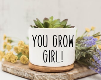 May include: White ceramic planter with a succulent plant and the text "YOU GROW GIRL!" printed in black. The planter is sitting on a wooden surface.