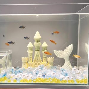 May include: An aquarium with a light green castle, white mermaid tail, and colorful gravel. Several small orange and blue fish swim in the clear water. The castle has a detailed design with turrets and a central tower.