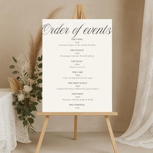 Può includere: Un cartello nuziale color crema su un cavalletto di legno mostra l'"Ordine degli eventi" in un'elegante scritta nera. Il cartello elenca la cerimonia, i brindisi, il banchetto, la torta, il primo ballo, la festa e l'addio.