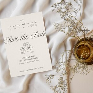 Pode incluir: Um convite de casamento creme com o texto "Save the Date" em escrita elegante. O cartão apresenta um calendário de maio, duas taças de champanhe e detalhes do casamento. Um arranjo floral decorativo e uma tigela dourada estão no fundo.