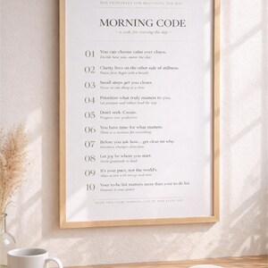 May include: A framed print with the text "MORNING CODE" and ten principles for starting the day. The print is in a light wood frame and is on a wooden desk with a coffee mug and an open book.