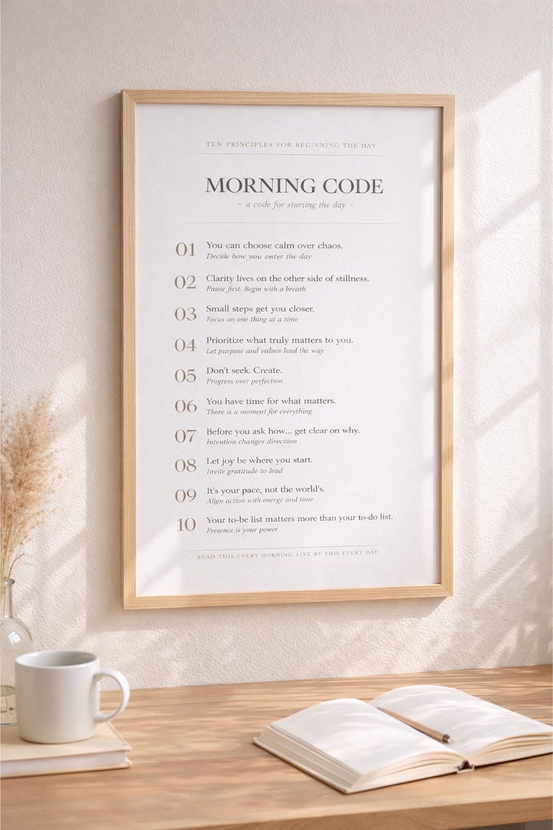May include: A framed print with the text "MORNING CODE" and ten principles for starting the day. The print is in a light wood frame and is on a wooden desk with a coffee mug and an open book.