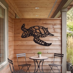 May include: A black metal wall art piece shaped like a sea turtle with a detailed shell pattern and flippers. The cutout design features open spaces within the body and limbs, creating a silhouette that is mounted flat against a wooden wall.