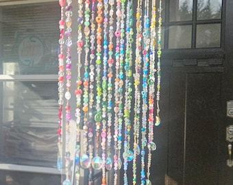 May include: A whimsical wind chime crafted from a weathered wooden branch, adorned with strands of colorful beads in various shapes and sizes. The strands hang down, interspersed with iridescent crescent moon charms. A small mushroom and greenery decorate the top.