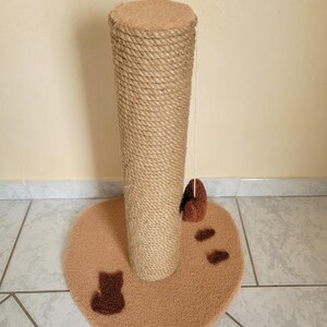 May include: A cat scratching post with a beige, heart-shaped base and a tall, rope-wrapped post. The base features brown cat silhouettes and paw prints. A dangling toy hangs from the top of the post. The post is designed for cats to scratch and play.