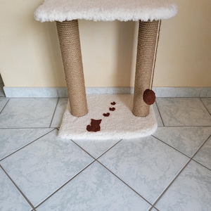 Peut inclure: Un petit arbre à chat avec un dessus et une base rectangulaires recouverts d'un matériau blanc et duveteux. Deux poteaux enveloppés de corde naturelle soutiennent le dessus. Une boule marron est suspendue en haut, et une silhouette de chat et des cœurs marron ornent la base.