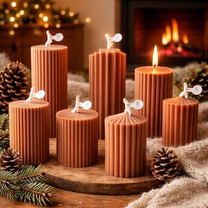 Peut inclure: Plusieurs bougies piliers marron avec des détails côtelés verticaux. Certaines bougies ont des étiquettes blanches attachées. Une bougie est allumée, avec une flamme. Les bougies sont disposées sur une surface en bois avec des pommes de pin et de la verdure.