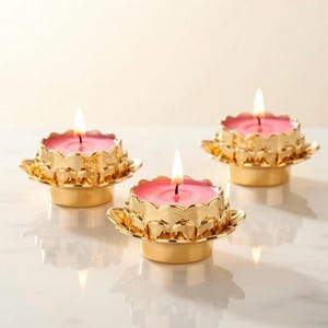 Peut inclure: Trois bougies allumées avec de la cire rose sont placées dans des bougeoirs dorés en forme de fleur de lotus. Les bougeoirs ont un design de pétales détaillé et reposent sur une surface blanche. L'ensemble est élégant.