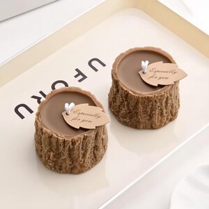 May include: Two brown candles shaped like tree stumps, each with a smooth top and textured sides. A small leaf-shaped tag with the text "Especially for you" is attached to each candle. The candles are displayed on a white tray.