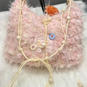 Op de afbeelding: Een zachte, roze handtas met een crèmekleurige riem en houten kralen. De tas heeft een knoopsluiting en decoratieve knopen. Een klein label met teddybeer illustraties en de tekst "A Gift For" is zichtbaar.