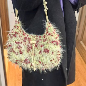 May include: A fuzzy, light green handbag with red accents. The bag has a crescent shape and a drawstring closure with a wooden toggle. The strap is made of a cream-colored cord with wooden beads.