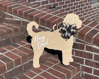 May include: A wooden dog-shaped sign with the words "Hey Dood" in white script. The sign has a black and white checkered bow and a string of white beads. The sign is light beige and is displayed on a brick wall.