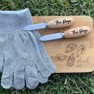 May include: A wooden cutting board with engraved oyster illustrations and the text "shuck em". Two oyster knives with wooden handles engraved with "The Rays" are next to the board. A gray cut-resistant glove is also in the image.