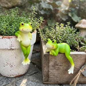 May include: Two green frog figurines. One frog is standing on a white pot, the other is lying on a wooden box. Both frogs have yellow eyes and white feet. The background includes green plants and a mushroom statue.