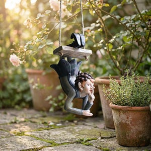 May include: A whimsical garden ornament featuring a fairy figure suspended from a wooden swing. The fairy has red hair, a thoughtful expression, and is dressed in a blue and gray outfit. The swing hangs from a rope, with a blurred background of plants and pots.