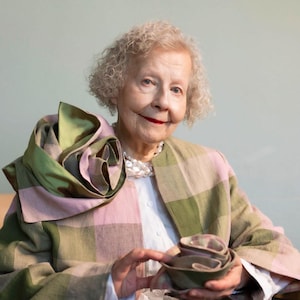 May include: An older person wearing a pink and green plaid jacket and a white shirt, holding a stack of small, green bowls. The jacket has a large, decorative scarf in shades of green and pink. A pearl necklace is also visible.