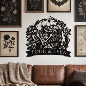 Op de afbeelding: Een zwart metalen wanddecoratie met een skeletkoppel dat elkaar omhelst binnen een cirkelvormig frame van rozen en bladeren. Onder de figuren staat een banner met de tekst TODD & LEEA in witte hoofdletters op een witte muur.