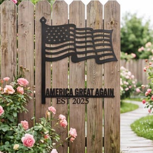 Puede incluir: Un letrero de metal negro con un diseño de bandera estadounidense está montado en una valla de madera. El letrero incluye el texto "AMERICA GREAT AGAIN EST 2025". La valla está rodeada de rosales rosas y una zona de césped.