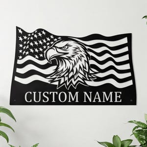May include: Black metal wall art featuring an American flag design with a detailed eagle head. The flag includes stars and stripes, with the text "CUSTOM NAME" below the eagle. This patriotic decor is suitable for indoor or outdoor display.