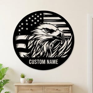 May include: A circular black and white wall sign featuring a detailed illustration of a bald eagle head in profile, layered over a stylized American flag design. The text CUSTOM NAME is printed in a bold, sans-serif font at the bottom center.