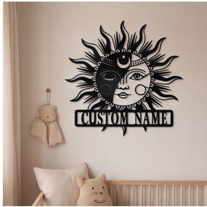 Pode incluir: Uma peça de arte de parede em metal preto com um design de sol e lua com um rosto central. Os raios de sol estendem-se para fora e uma faixa retangular na parte inferior contém o texto CUSTOM NAME numa fonte serifada em negrito.