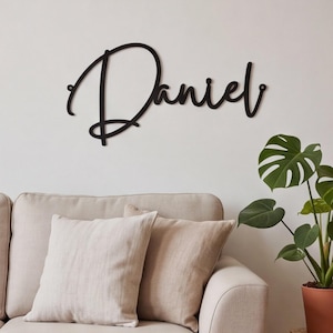 May include: A black metal wall sign featuring the name Daniel written in a continuous cursive script font. The sign has two small circular mounting holes, one on the far left and one on the far right, for attachment to a flat surface.
