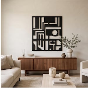 May include: A square black wall art piece featuring a geometric cutout design with straight lines and a circular opening. The artwork is mounted on a plain wall above a long wooden sideboard, which holds a vase with green branches and a small bowl.