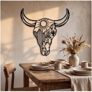 May include: A black metal wall art piece shaped like a bull skull, featuring a cutout design of a desert landscape with a sun, tall saguaro cacti, and prickly pear cacti. The intricate silhouette is mounted on a plain wall above a dining table.