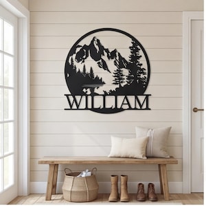 May include: A circular black metal wall sign featuring a laser-cut mountain range, pine trees, and a lake scene. The name WILLIAM is cut out in capital letters across the bottom section of the circular frame, integrated into the design.