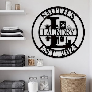 May include: A circular black metal wall sign featuring the text SMITH'S LAUNDRY EST. 2024. The design includes a large letter L, a washing machine icon, a laundry basket, a detergent bottle, a clothes hanger, and silhouettes of various garments.