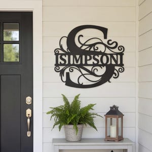 May include: A black metal wall sign featuring a large capital letter S with intricate scrollwork and the name SIMPSON cut out in the center. The sign is mounted on a white horizontal paneled wall next to a black door and a potted fern.