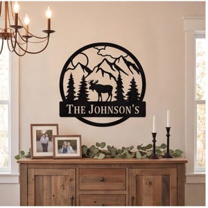 Könnte beinhalten: Ein rundes Wandbild aus schwarzem Metall mit der Silhouette eines Elchs in einem Wald, ergänzt durch Berge und eine Sonne im Hintergrund. Ein horizontaler Balken am unteren Rand zeigt den Text THE JOHNSON'S in weißer Serifenschrift.