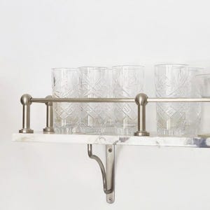 Op de afbeelding: Een marmeren plank met een metalen reling en beugels, met daarin verschillende heldere, bedrukte drinkglazen. De glazen hebben een geslepen ontwerp. De plank is aan een witte muur bevestigd.