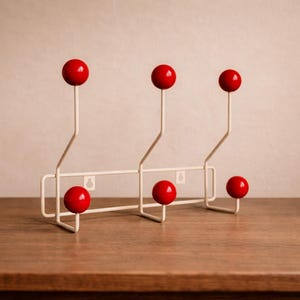 以下が含まれることがあります： 6つの赤いボール型のノブが付いた白い金属製のコートラック。ラックには上部に3つ、下部に3つのノブがあります。木製のテーブルの上にあります。背景は無地でニュートラルな色です。