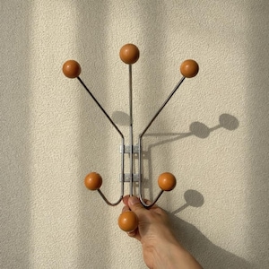Könnte beinhalten: Ein Garderobenständer im Mid-Century-Stil mit sechs holzfarbenen, kugelrunden Haken in einem warmen, hellbraunen Farbton. Die Haken sind an einem silberfarbenen Metallrahmen befestigt. Der Garderobenständer ist an einer strukturierten, cremefarbenen Wand befestigt.