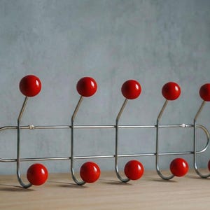 Könnte beinhalten: Moderner Kleiderhaken mit silbernem Metallrahmen und sechs leuchtend roten kugelförmigen Haken. Die Haken sind symmetrisch angeordnet, drei oben und drei unten. Der Haken steht auf einer hellen Holzoberfläche. Ca. 40 cm breit.